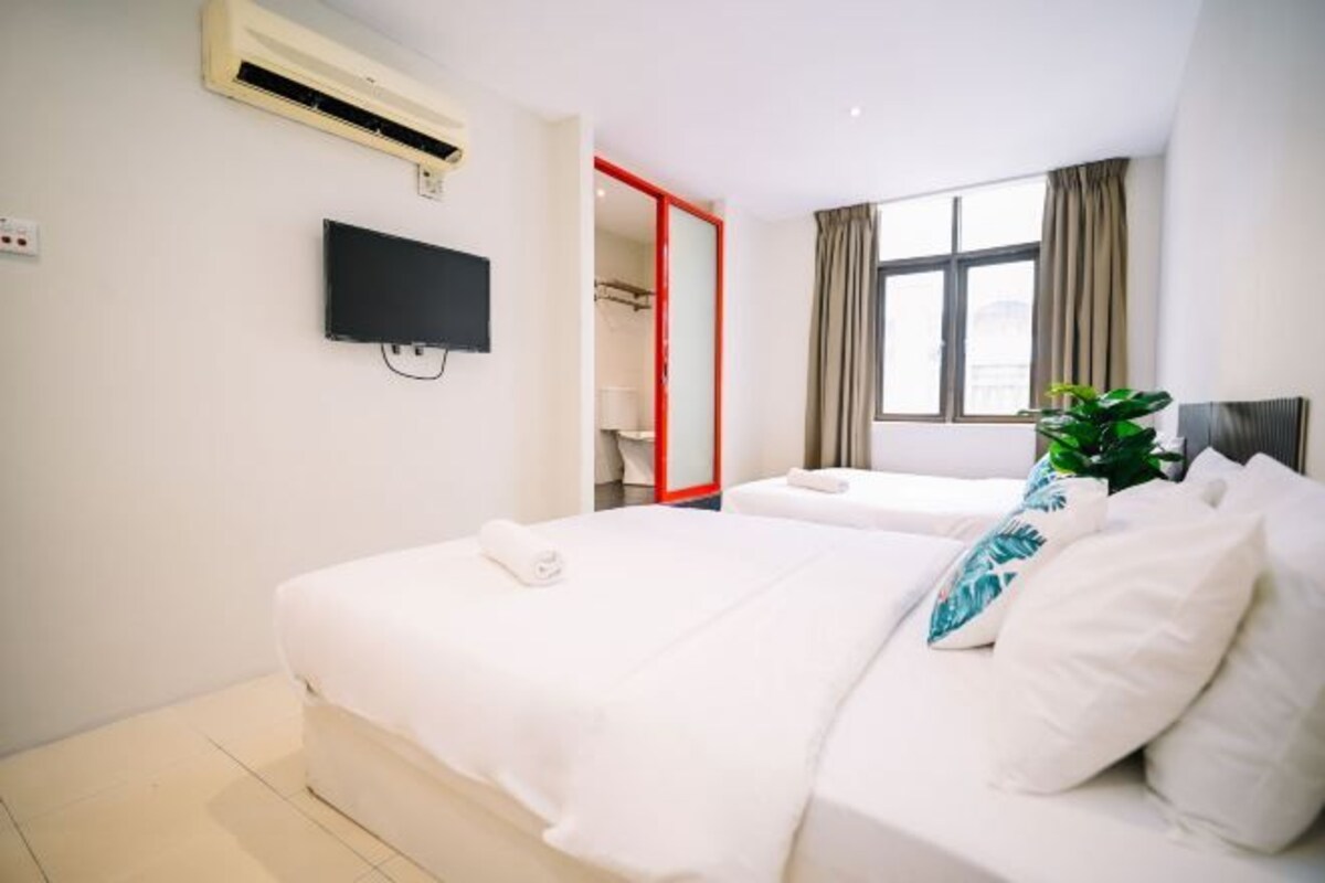 A hotel room with two beds, a TV, an air conditioning unit, a window with curtains, and a plant.