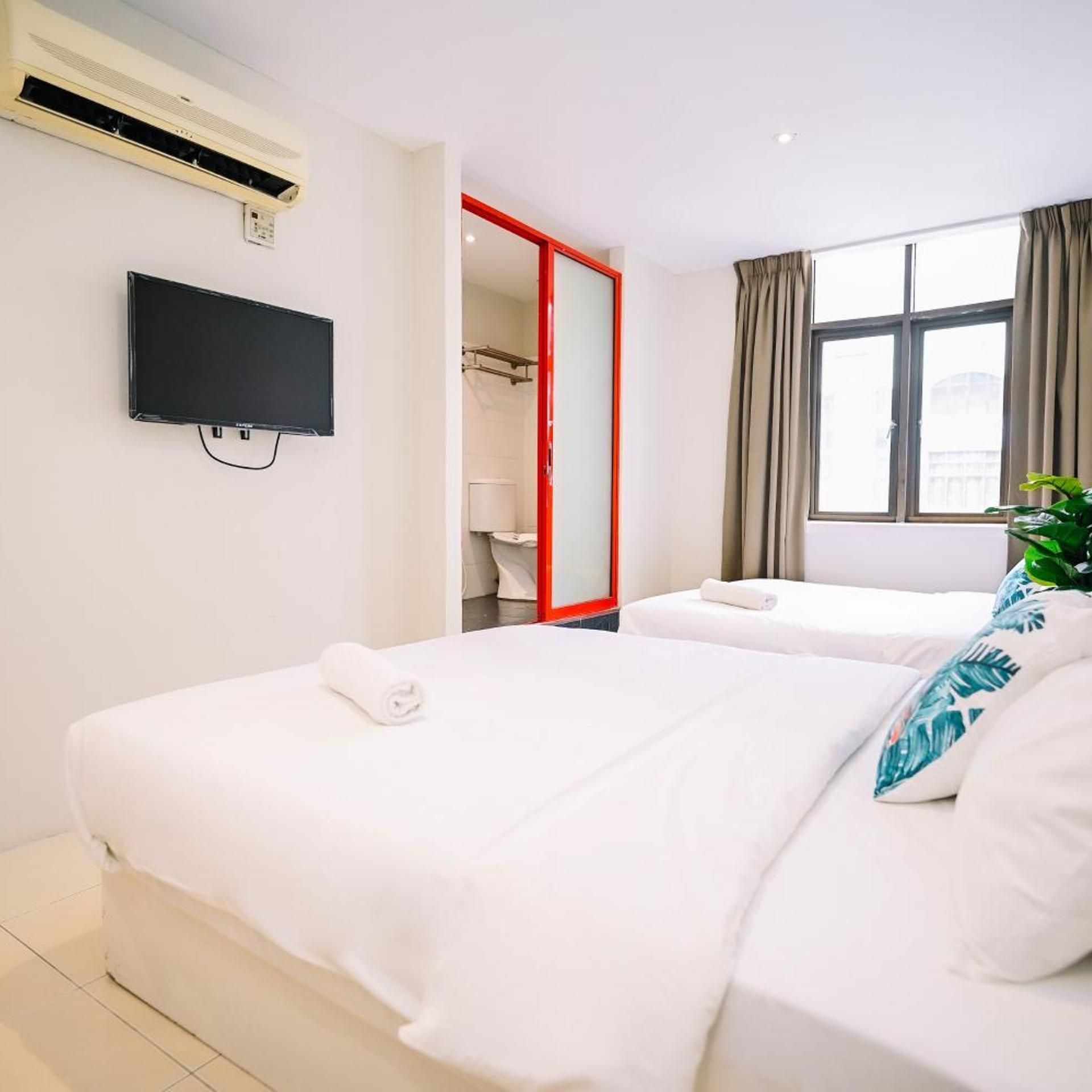 A hotel room with two beds, a TV, a window with curtains, and a red-framed door leading to a bathroom.