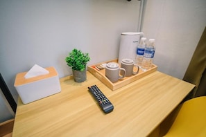 Ein Hotelzimmer mit einem Holztisch, einer Fernbedienung, einer Taschentuchbox, einem kleinen Topfpflanzen, einem Tablett mit einer Tasse, einer Wasserflasche und einer Kaffeemaschine.