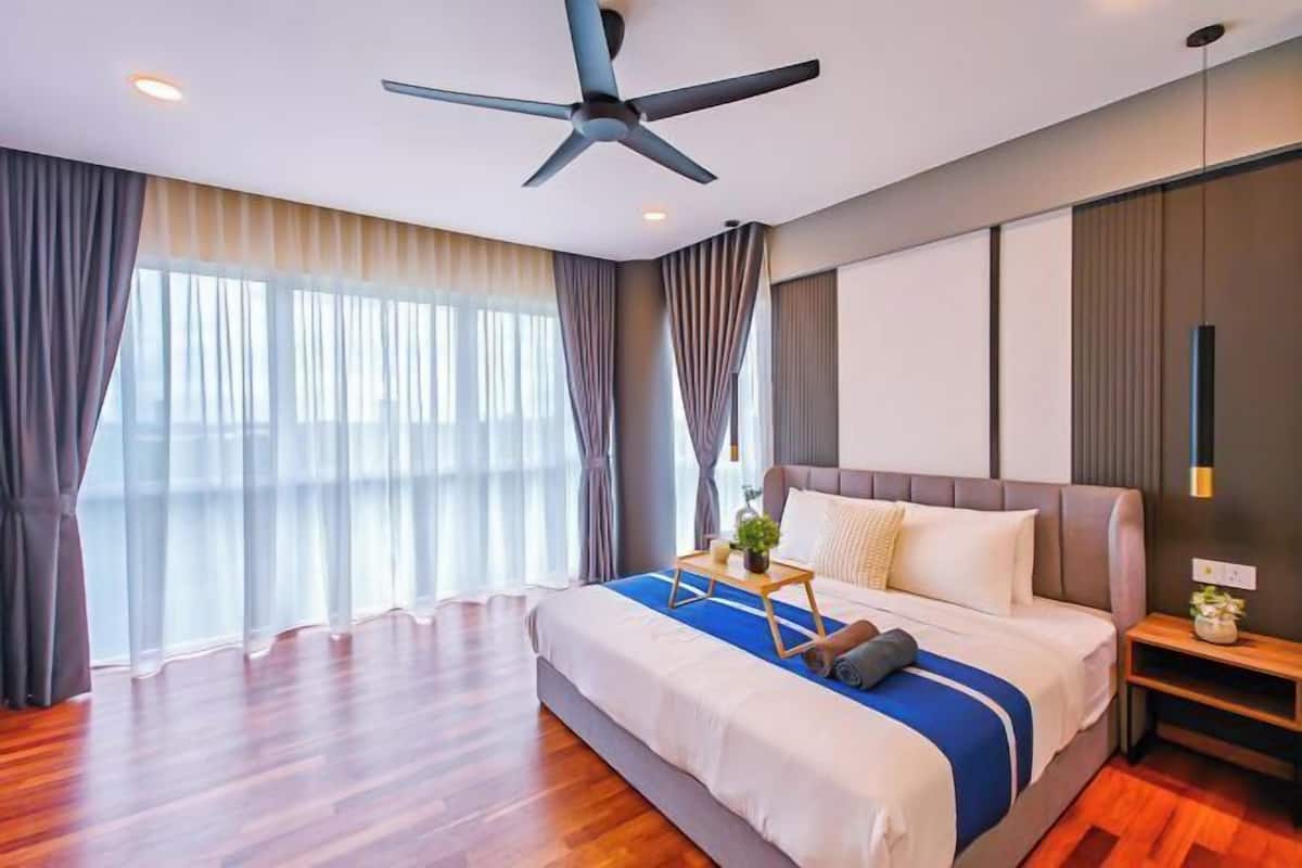 Una habitación de hotel moderna con una cama grande, un ventilador de techo, una mesita de noche con lámpara y vistas a la ciudad a través de amplias ventanas.
