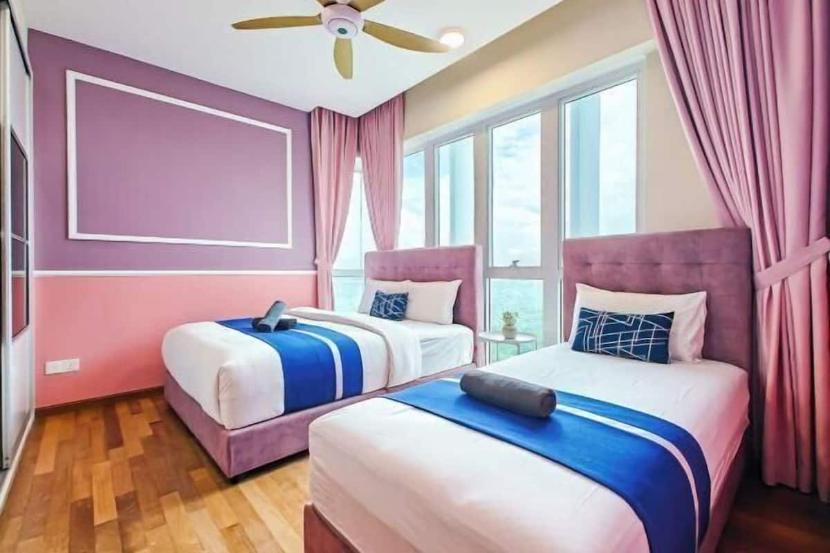 Habitación de hotel con dos camas, ventilador de techo, ventanas amplias y cortinas rosas.