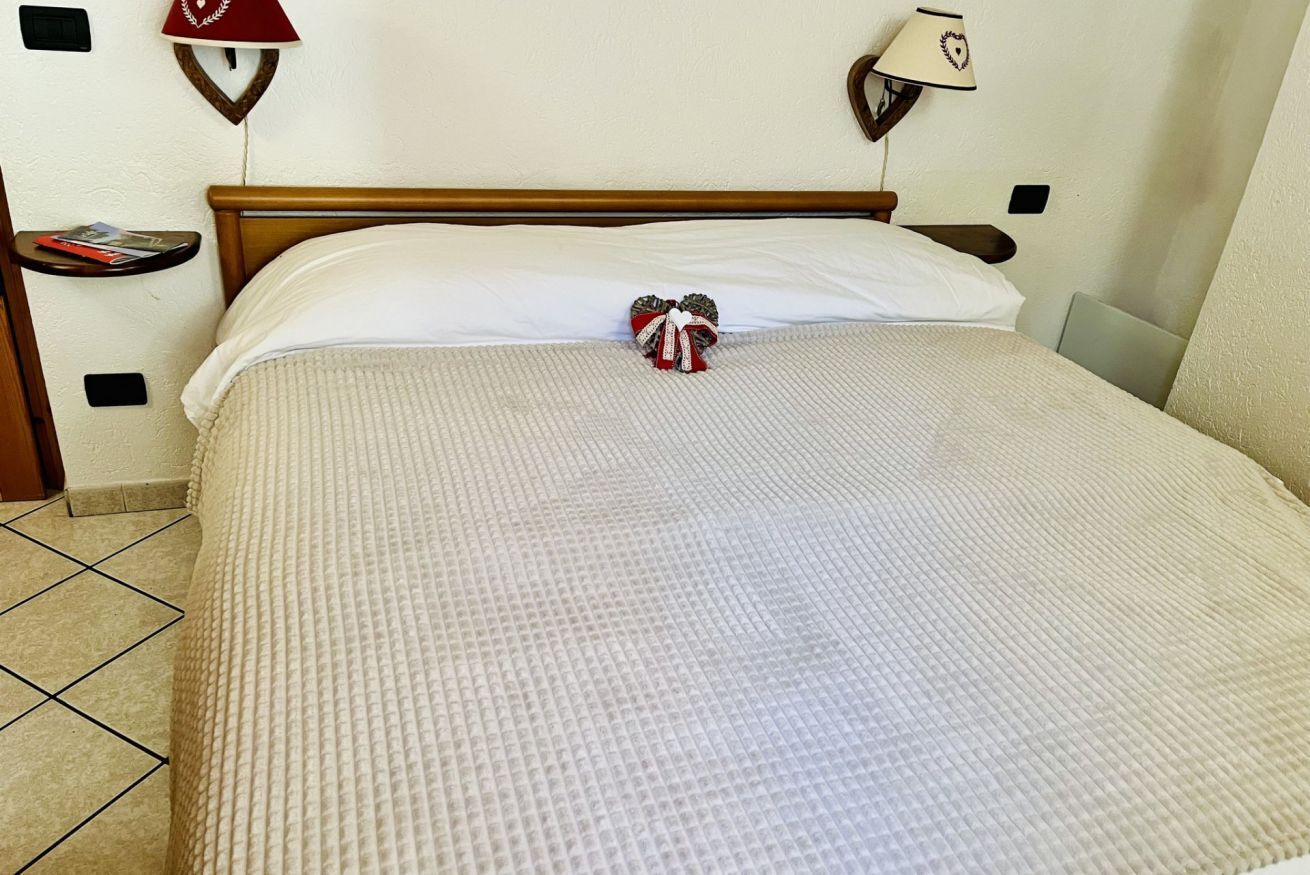Una cama bien hecha con un almohadón blanco y un lazo decorativo, cabecera de madera, una mesita de noche con una lámpara, y un estante pequeño con un libro.
