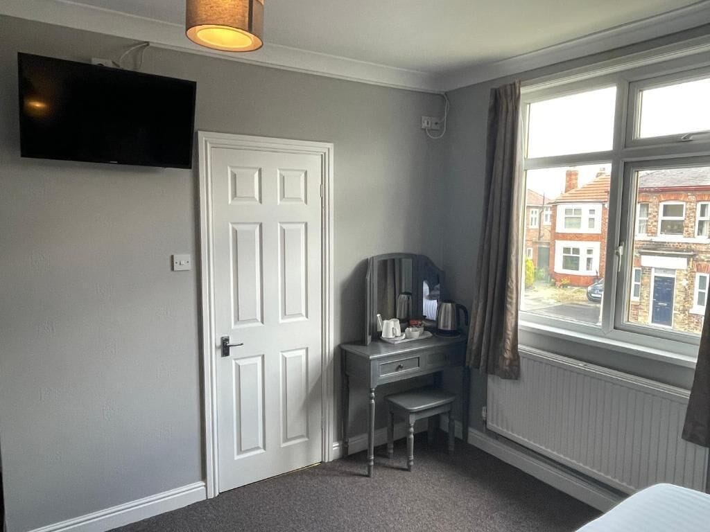 Ein Hotelzimmer mit weißer Tür, einem Schreibtisch mit Stuhl, einem an der Wand befestigten Fernseher und einem Fenster mit Vorhängen.