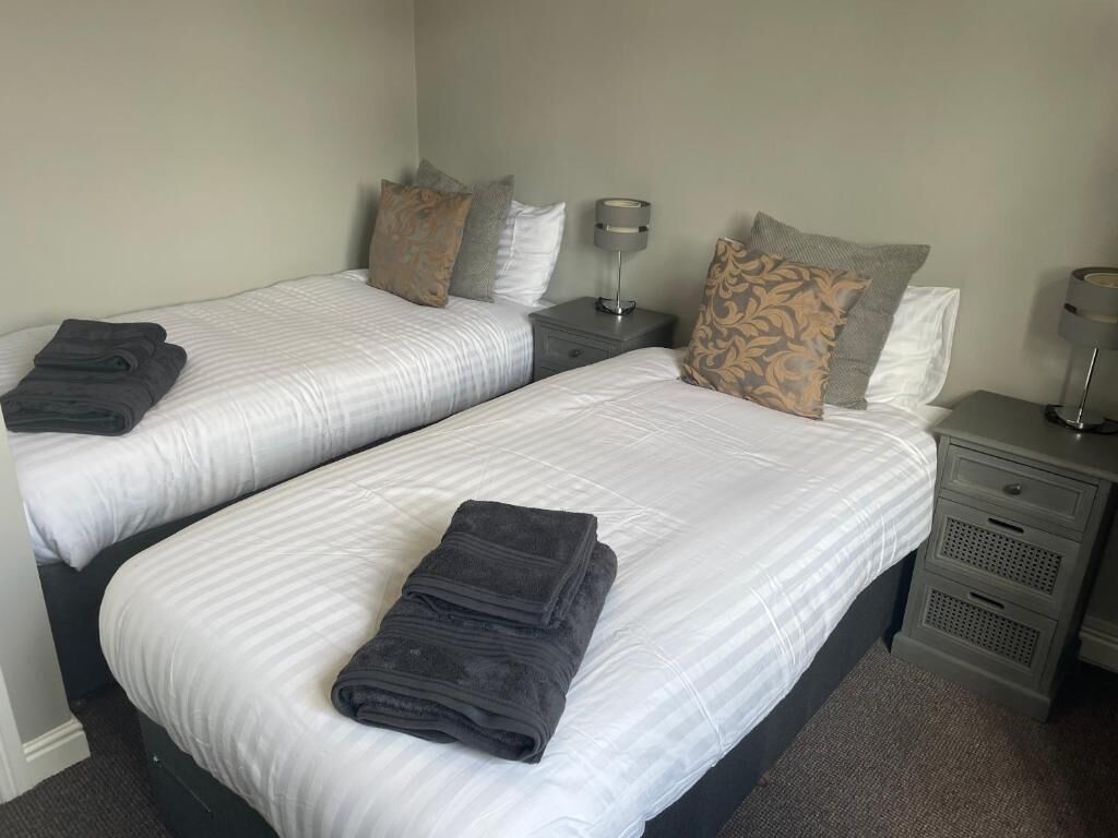 Une chambre d’hôtel avec deux lits, chacun recouvert d’une courtepointe blanche et agrémenté de serviettes sombres. On y trouve des tables de chevet avec des lampes et un petit tiroir.