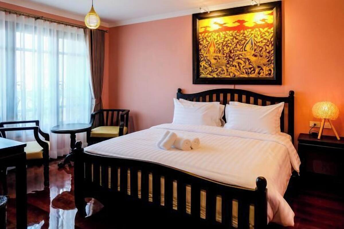 A neatly made bed with pillows and a rolled towel, a wooden nightstand with a lamp, a chair, and a table with a lamp.