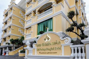 Un hotel de varios pisos con fachada amarilla, un cartel que indica que es el Sirikit Heritage Bangkok Hotel, y una escalera que conduce a la entrada.