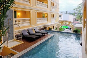 Un área de piscina con sillones reclinables, una terraza de madera y un edificio con balcones y un letrero.
