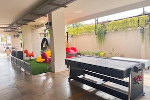 Una mesa de billar en una sala con zona de césped artificial verde, un tobogán rojo y una pared blanca con plantas trepadoras verdes.