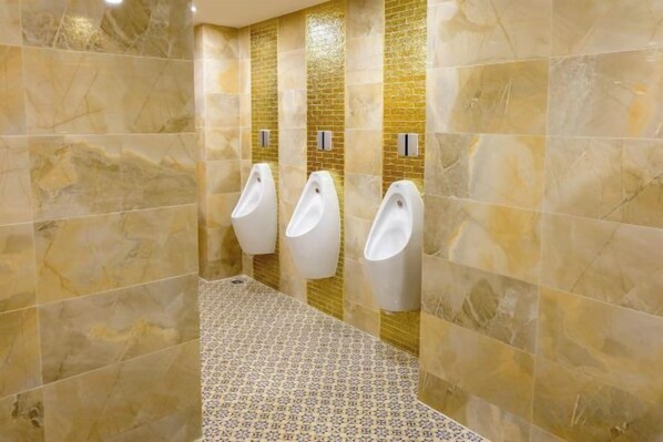 Des toilettes comprenant trois urinoirs, des murs en marbre et des carreaux de sol à motifs.