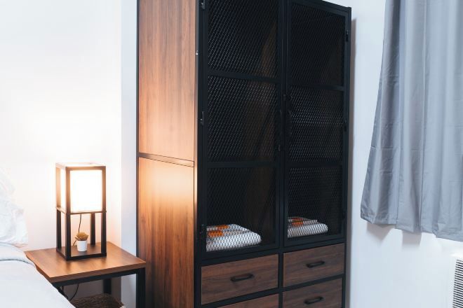 Un dormitorio con una mesita de noche de madera, una lámpara de noche, un armario con frente de malla y una cortina gris.