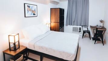 Un dormitorio con cama, mesita de noche con lámpara, armario, escritorio con sillas y ventana con cortinas.
