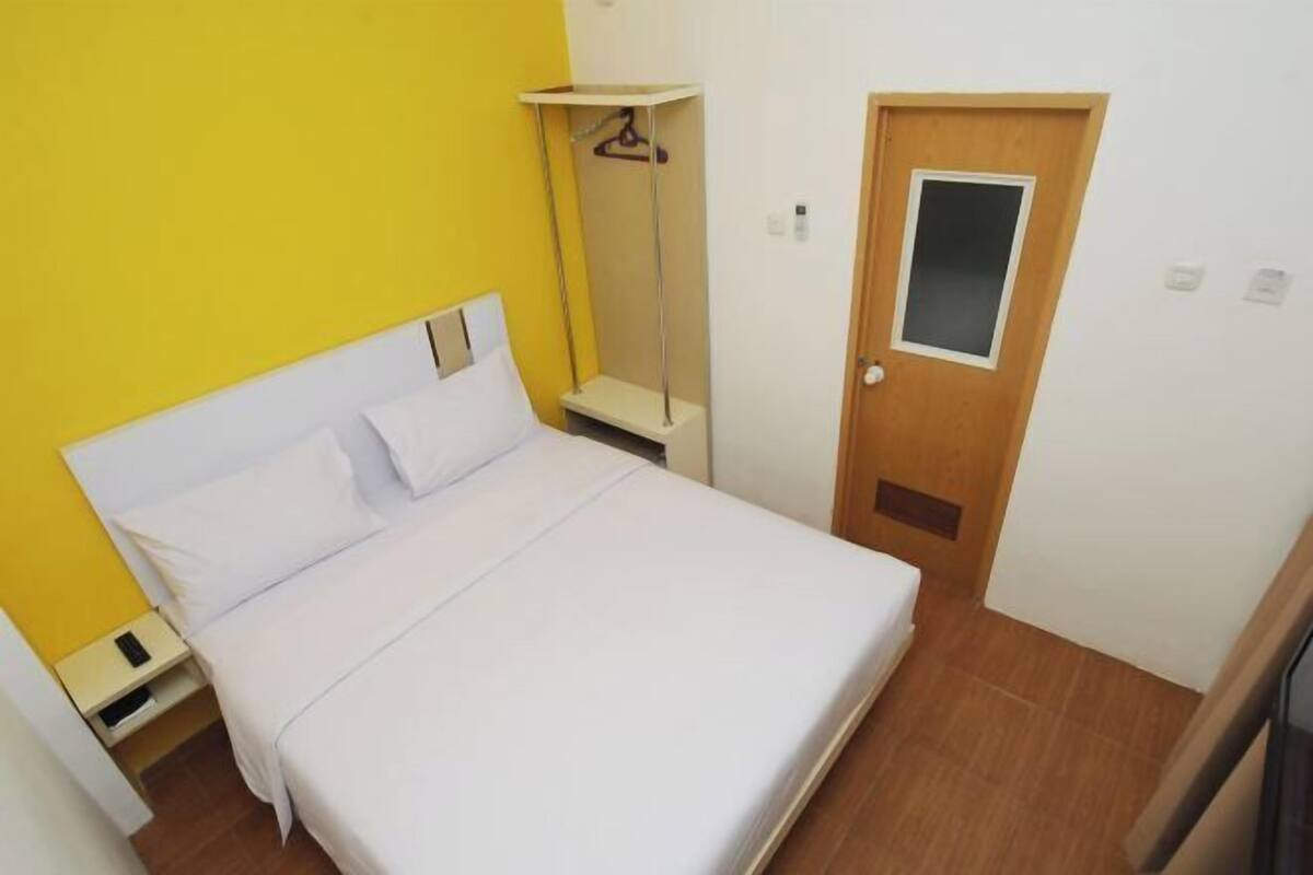 Un lit simple avec du linge de lit blanc, une porte en bois et une petite étagère avec un cintre.