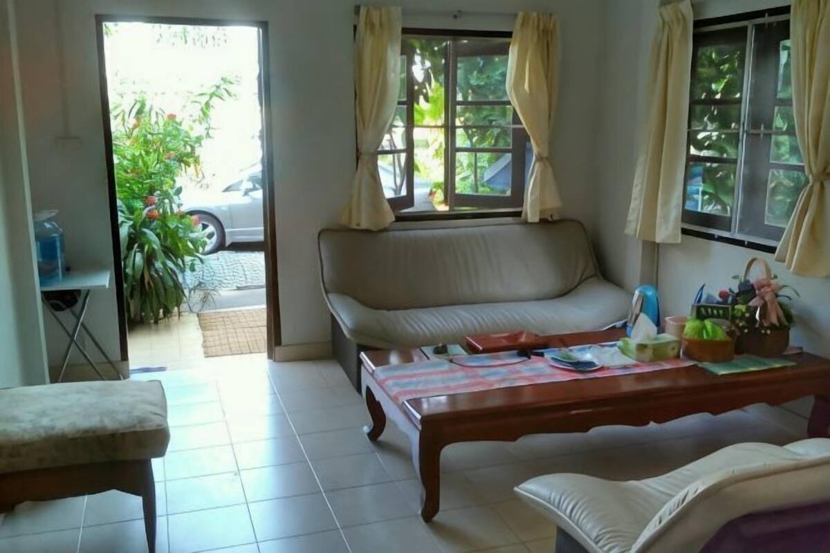 A living room with a sofa, a coffee table, and a chair. There are windows with curtains, a door leading outside, and a small table with items on it.