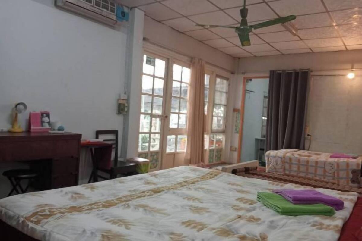 A room with a bed, a desk, and a chair. There is a window with curtains, a ceiling fan, and a door.