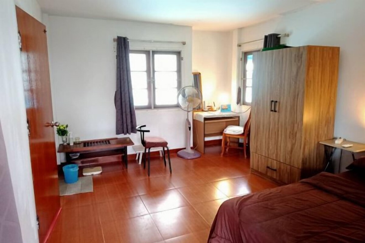 A bedroom with a bed, a desk, a chair, a wardrobe, a fan, and a window with curtains.