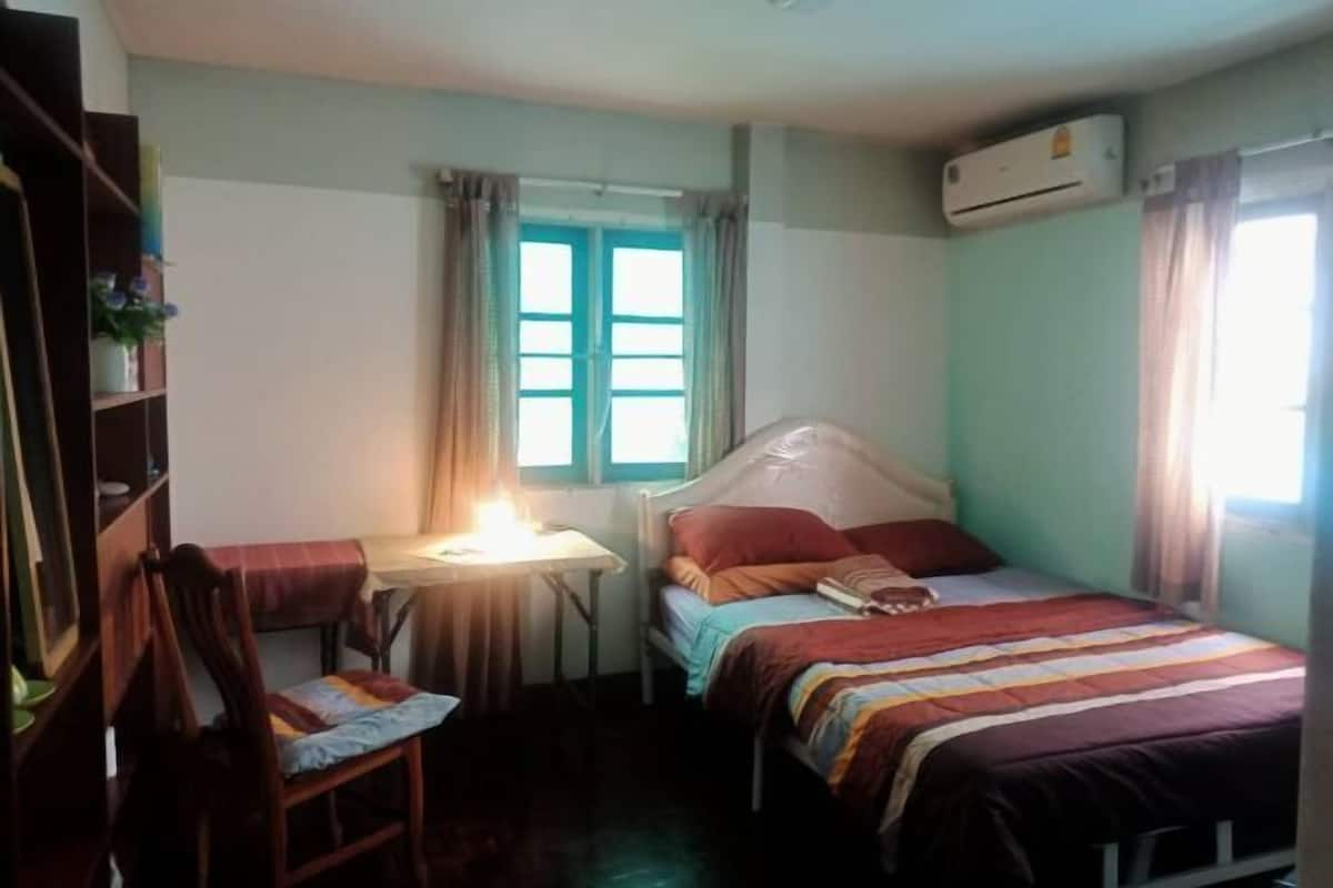 A bedroom with a bed, a desk, a chair, a window with curtains, and an air conditioning unit.