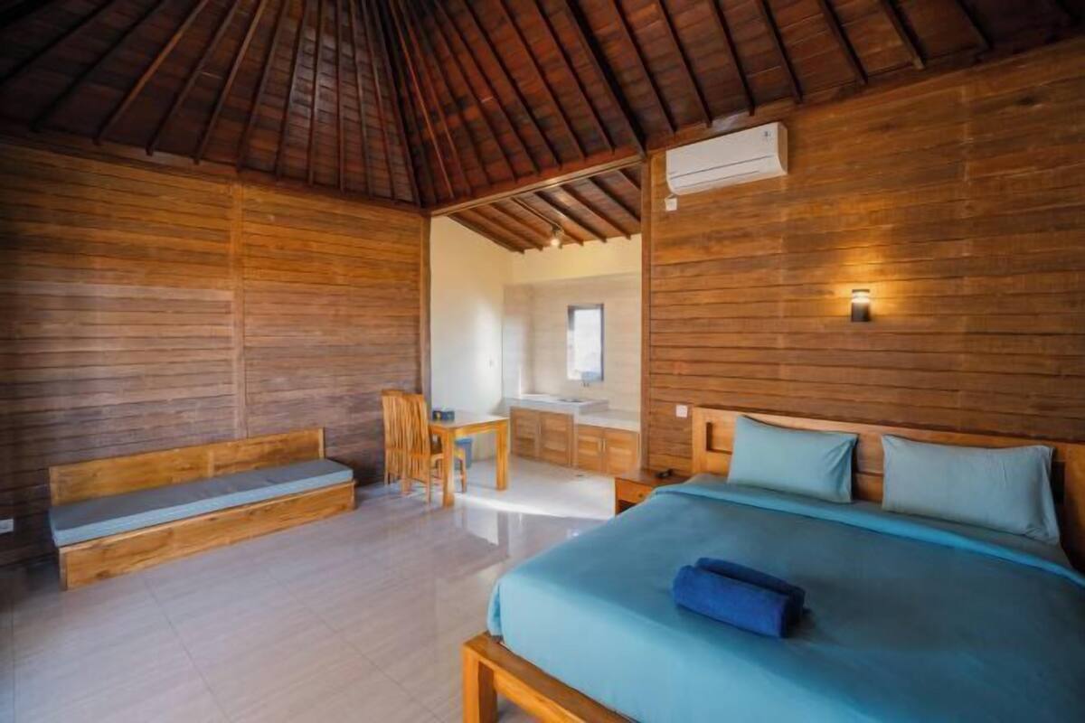A wooden room with a bed, a bench, a desk, and a bathroom visible through an open door.