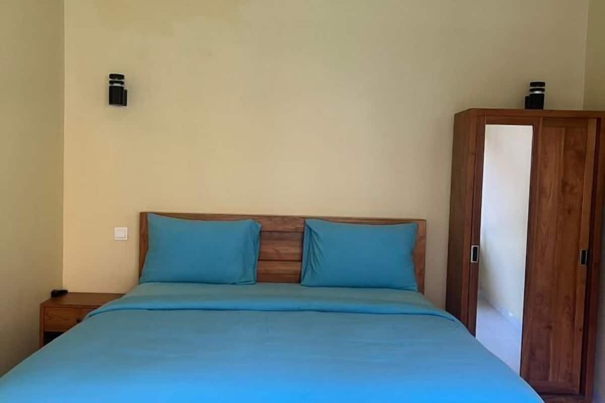A bedroom with a wooden headboard, two blue pillows, a wooden nightstand, and a wardrobe with a mirror.