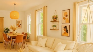 Una sala de estar moderna con un sofá blanco, una silla colgante, una zona de comedor con mesa y sillas, y ventanas amplias que dejan entrar luz natural.