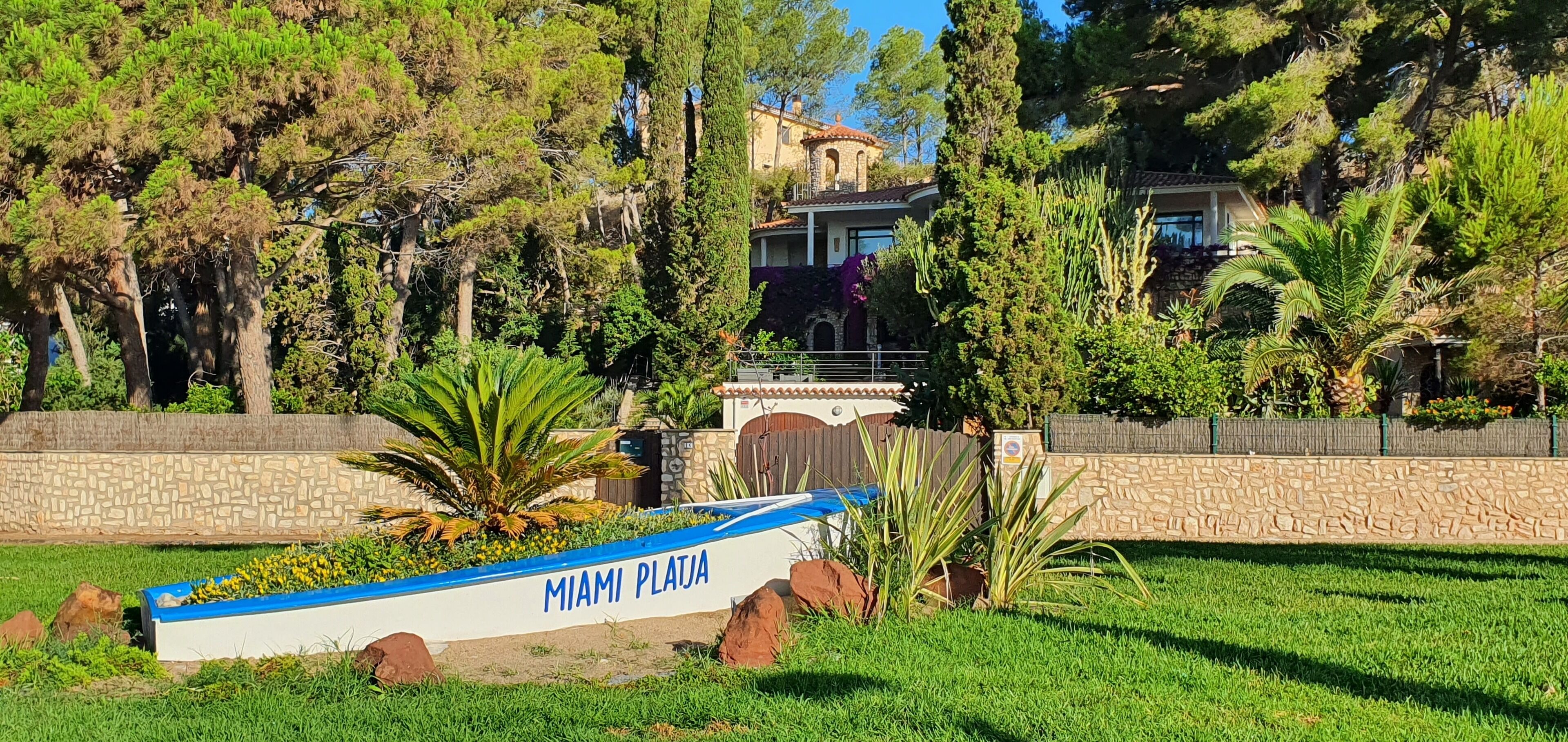 Un letrero que dice 'MIAMI PLAJA' frente a una casa rodeada de árboles y un muro de piedra.