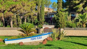 Placa com a inscrição 'MIAMI PLAJA' em frente a uma casa rodeada por árvores e um muro de pedra.