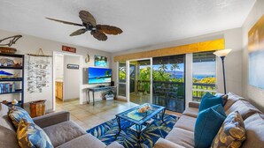 A living room with a sofa, coffee table, and a ceiling fan. There is a TV and a window with a view of palm trees. - Ocean views, 2BR retreat, Less than 5 Minutesʻ to the Beach - Kaanapali Plantation 20 (Lahaina)