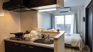 A compact hotel room with a kitchenette, a bed, a window with curtains, and a small bedside table. - Bainan Service Apartment (Osaka)