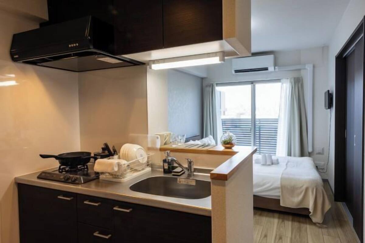A compact hotel room with a kitchenette, a bed, a window with curtains, and a small bedside table.