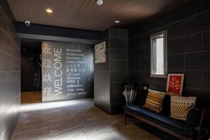 A dark-toned interior with a reception area featuring a welcome sign in multiple languages, a bench with cushions, and a window providing natural light. - Bainan Service Apartment (Osaka)