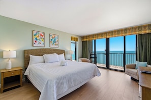 A hotel room with a large bed, two bedside lamps, a chair, a desk, and a view of the sea.