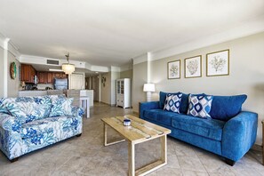 Un salon avec un canapé bleu, un canapé à motifs, une table basse en bois et, au fond, une cuisine.