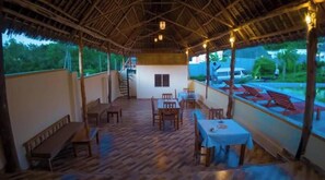 An outdoor dining area with wooden tables and benches, a thatched roof, and a view of a pool and greenery.