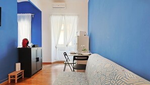 A room with a blue wall, a wooden floor, a sofa, a desk with a chair, a small cabinet, and a window with curtains.
