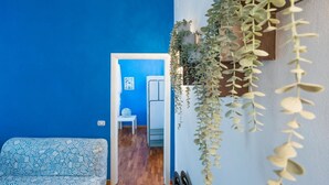 A room with a blue wall, a patterned sofa, a hanging plant, and a staircase leading to another room.