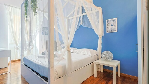 A room with a bed under a white canopy, a small bedside table with a decorative sphere, and a blue wall with a framed picture.