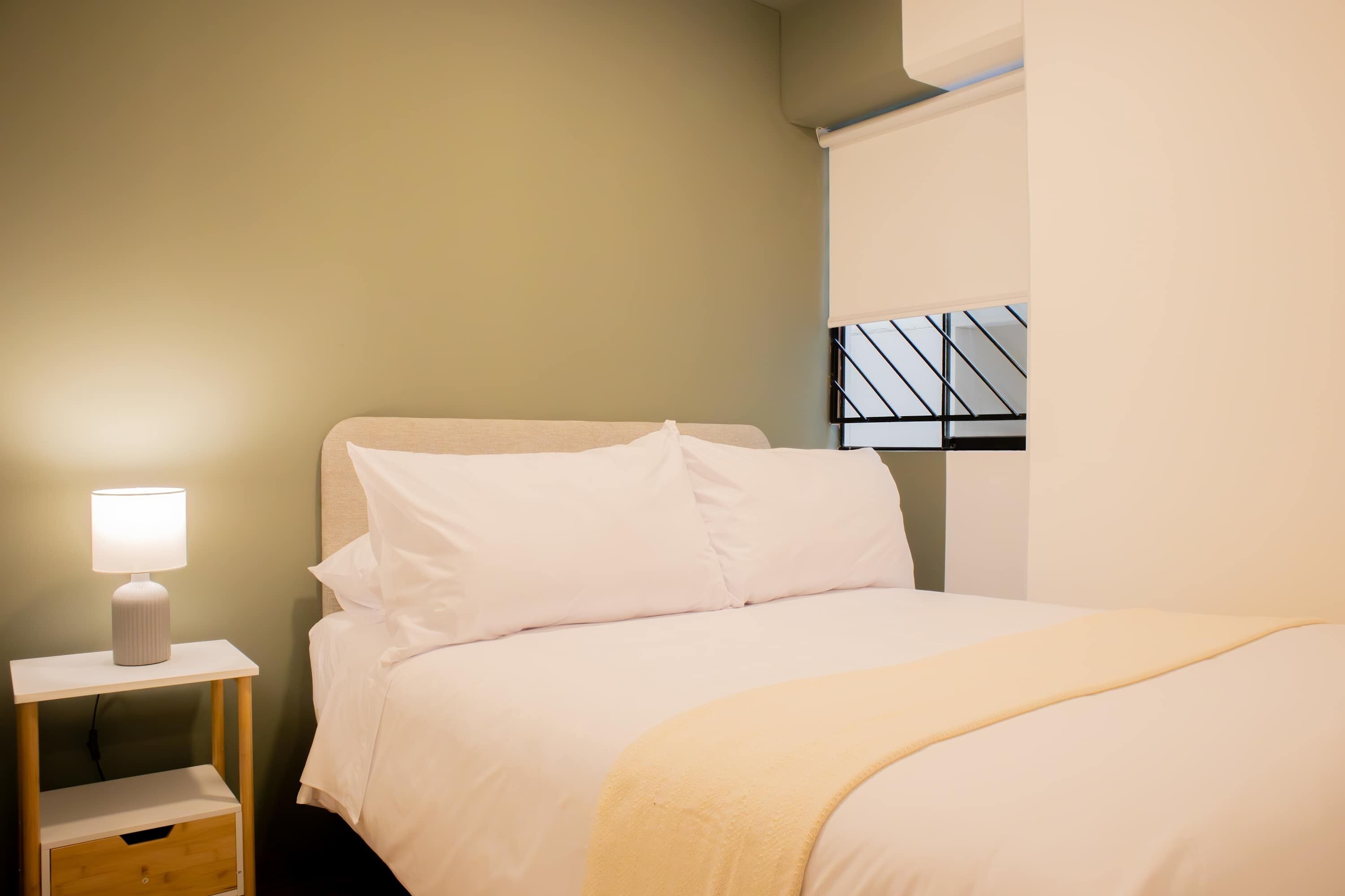 A neatly made bed with white bedding and a yellow throw blanket, a bedside table with a lamp, and a window with a black metal frame.