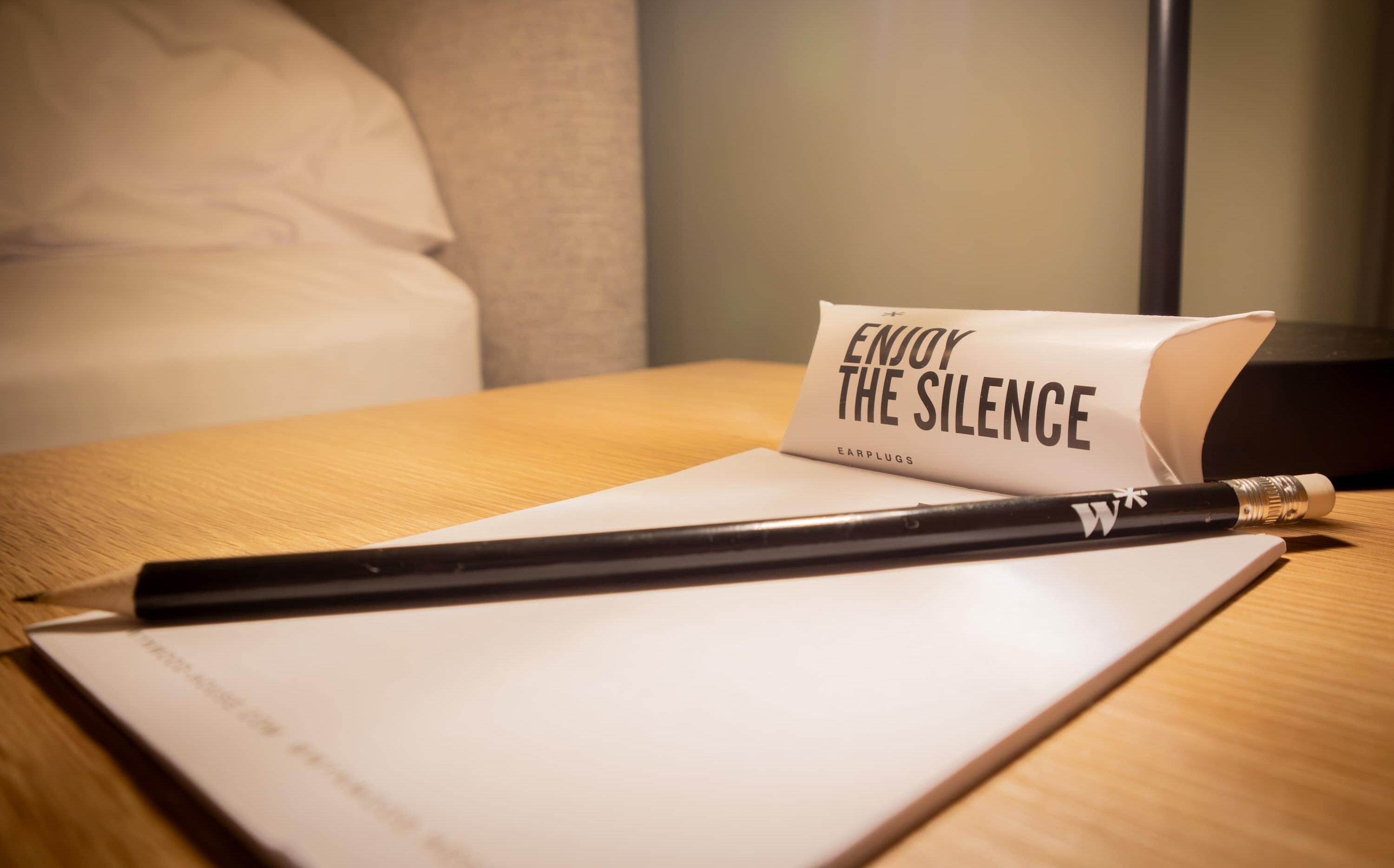 A notebook with a pencil on a wooden table, a sign that reads "ENJOY THE SILENCE" and the brand "EARPLUGS".