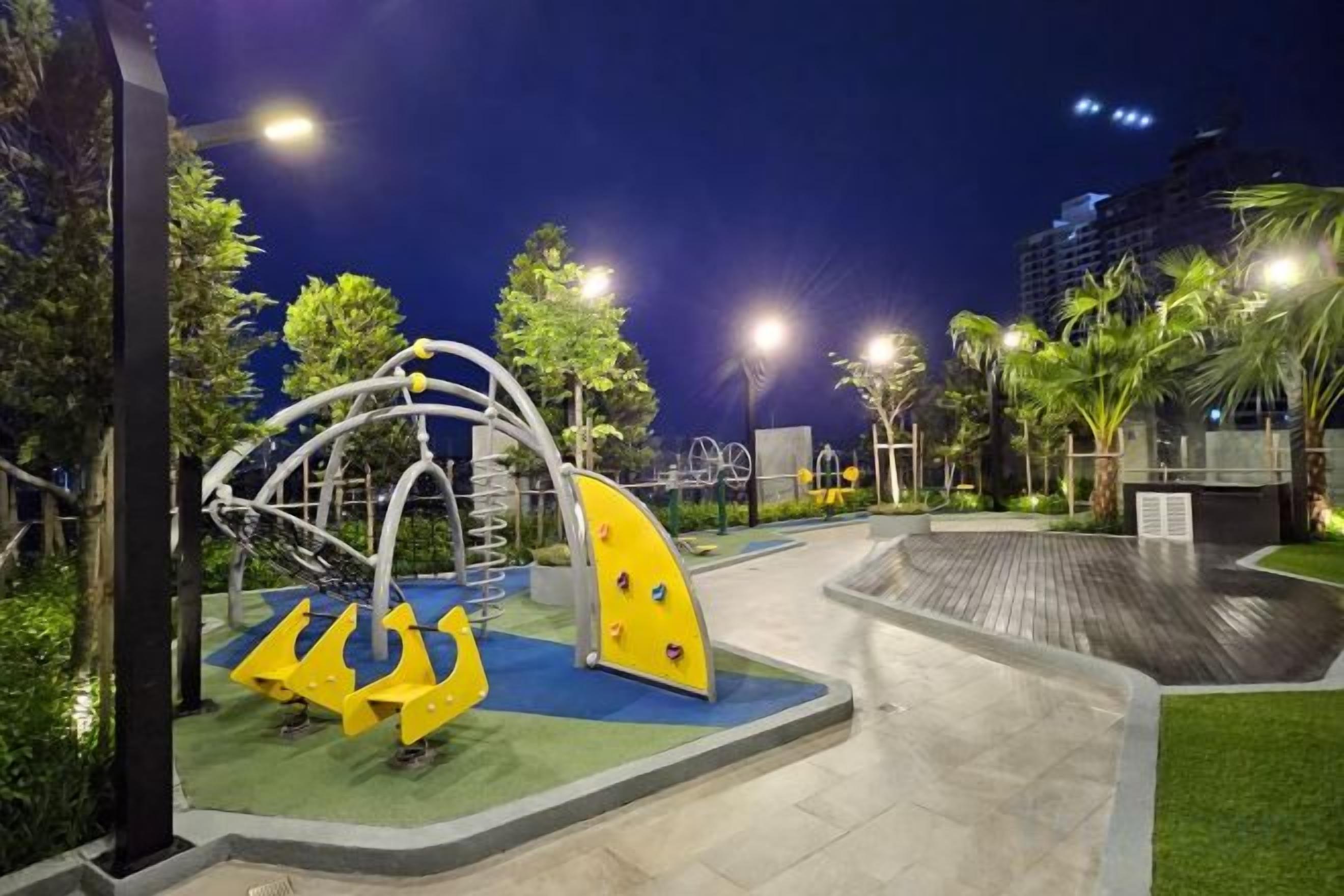 An outdoor playground with climbing structures and slides, surrounded by greenery and a paved walkway.