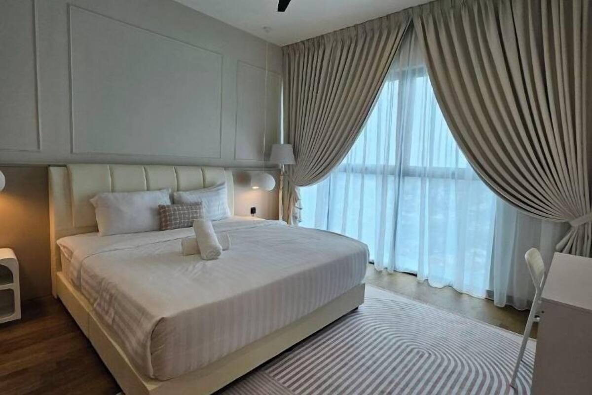 A bedroom with a large bed, a bedside table, a lamp, and a view of the outdoors through sheer curtains.