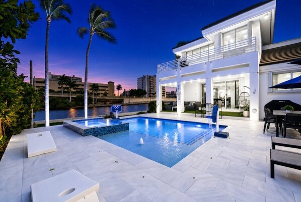 A modern outdoor pool area with a two-story building, palm trees, and a view of the water.
