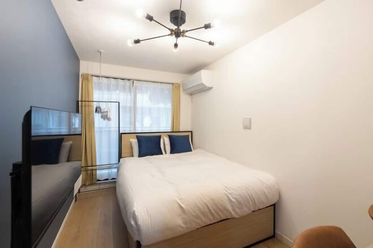 A bedroom with a bed, bedside tables, a desk, a chair, a ceiling light fixture, and a window with curtains.