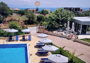 Ein Poolbereich mit Liegestühlen und Sonnenschirmen, umgeben von einer Stegterrasse und Grünpflanzen, mit Blick auf eine Küstenlandschaft.