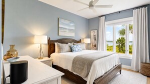 A bedroom with a bed, bedside lamps, a nightstand, a ceiling fan, a window with curtains, and a view of the outdoors. - 1906 South Beach Club - Just a Few Steps from Pool Days and Beach Waves! (Hilton Head Island)