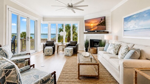 A spacious living room with a large window, a ceiling fan, a fireplace, and a television mounted on the wall. - 1906 South Beach Club - Just a Few Steps from Pool Days and Beach Waves! (Hilton Head Island)