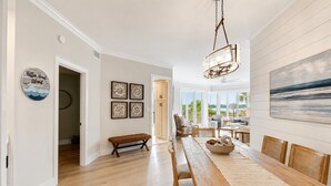 A spacious dining area with a long wooden table, wicker chairs, and a bench. There are framed artworks on the walls and a view of the outdoors through large windows. - 1906 South Beach Club - Just a Few Steps from Pool Days and Beach Waves! (Hilton Head Island)