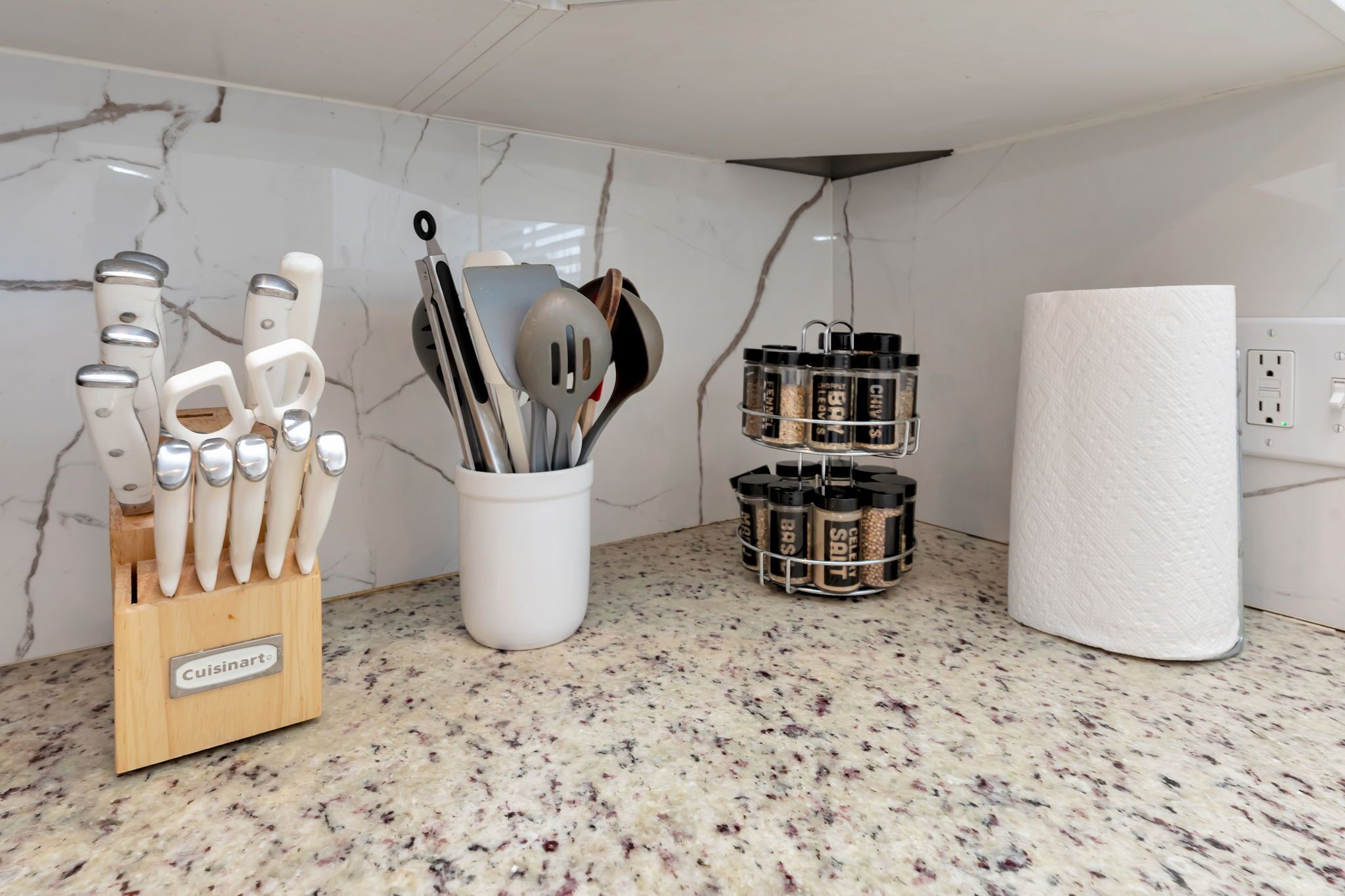 Cozinha com painel de fundo em mármore, porta-facas, suporte para utensílios, potes de tempero e rolo de papel toalha.