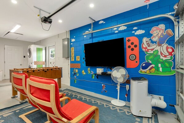 Una habitación con una amplia televisión de pantalla plana, una mesa de fútbol de mesa y una pared con temática de Nintendo.
