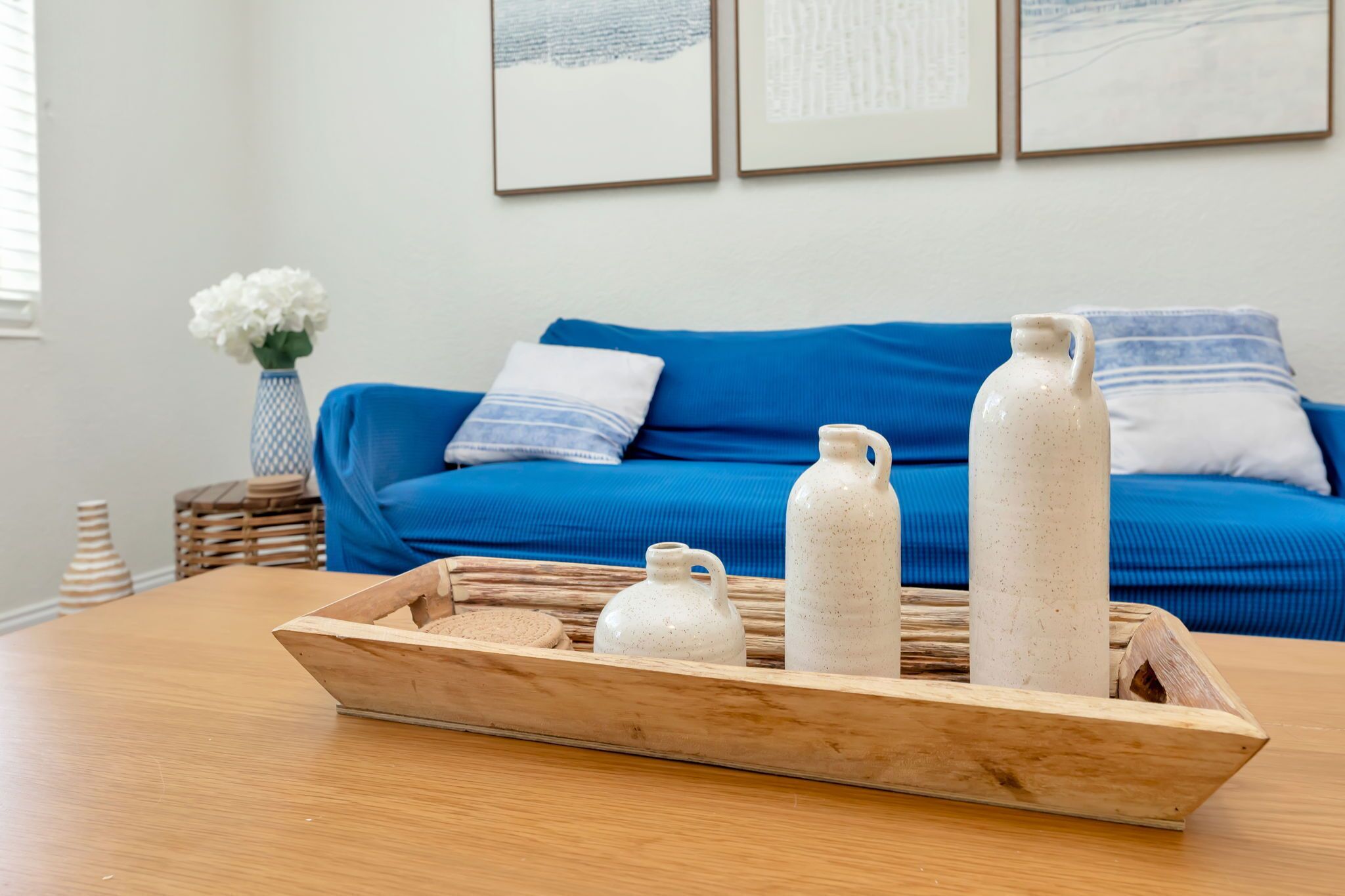 Un plateau en bois sur lequel reposent trois pichets en céramique blanche, le tout posé sur une table en bois. En arrière-plan, on aperçoit un canapé bleu orné de coussins à motifs et un vase rempli de fleurs blanches.
