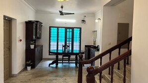 A spacious hallway with a marble floor, a staircase with wooden balusters, a ceiling fan, and a chandelier. - BLISS STAYS (Kanayannur)
