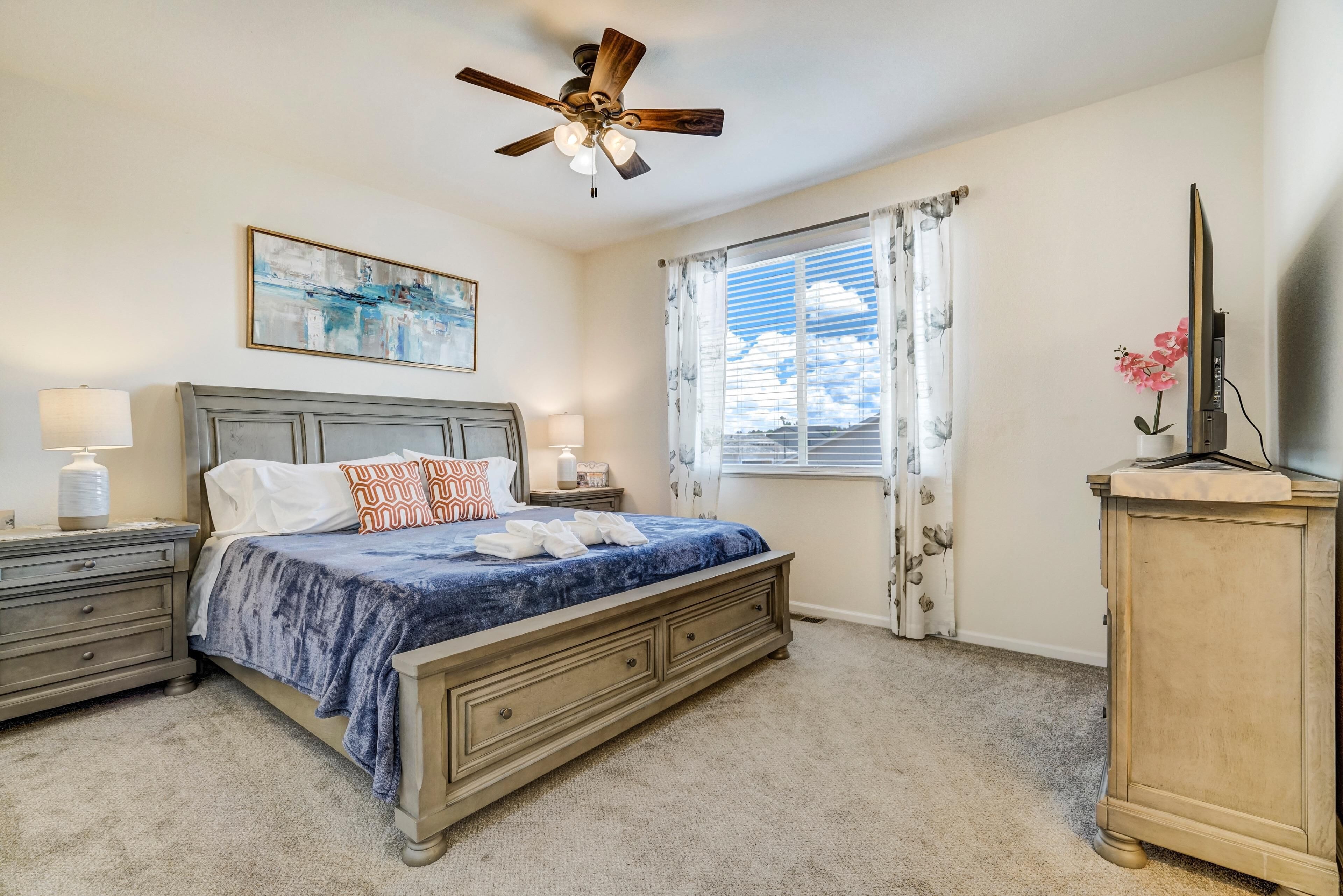 A bedroom with a bed, nightstands, a ceiling fan, a window with curtains, and a TV on a stand.